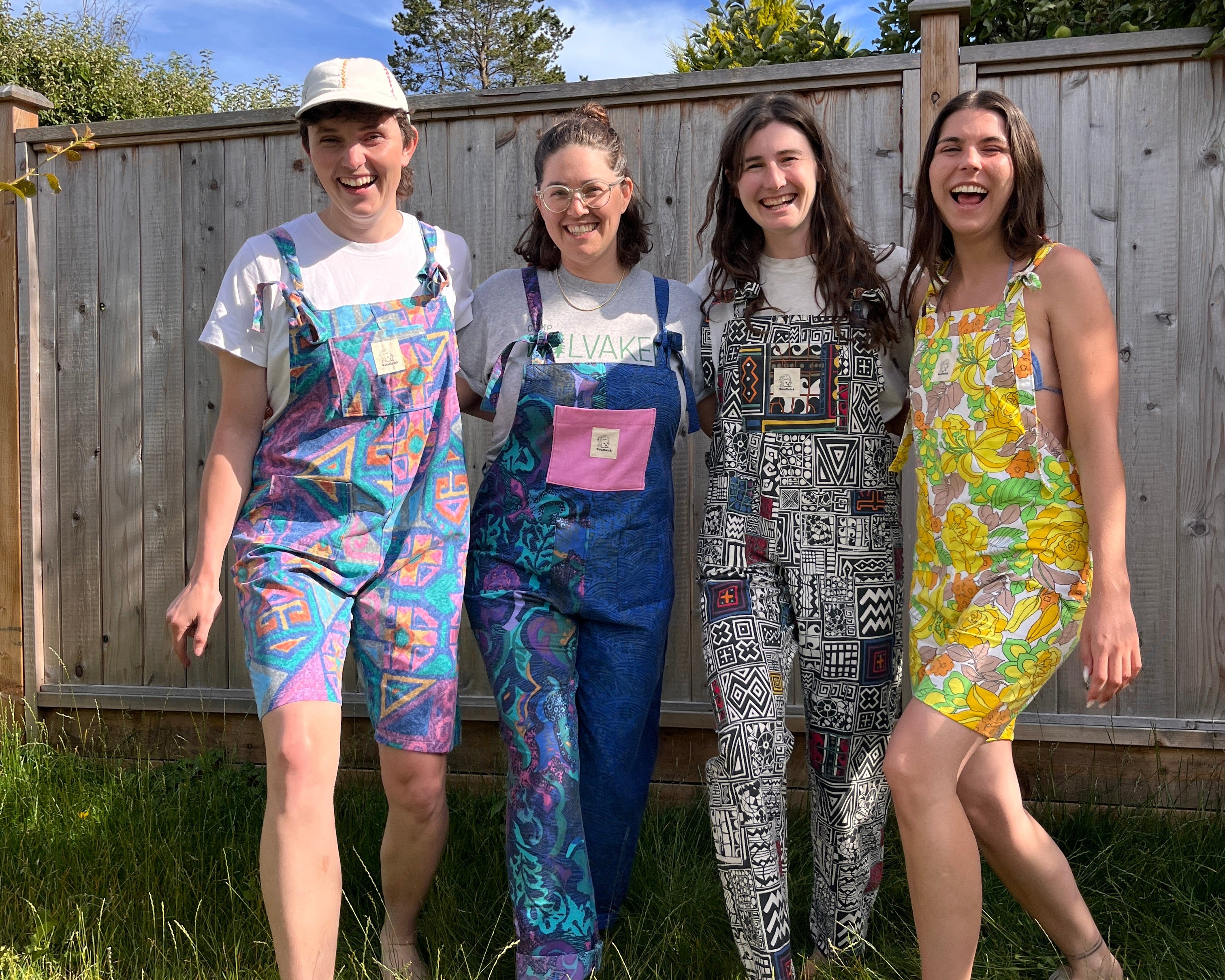 Four women standing outdoors in a backyard wearing colorful patterned jumpsuits and casual outfits, smiling and posing in front of a wooden fence under a blue sky.