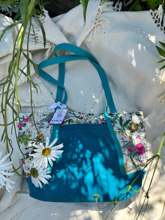 A handmade fabric tote bag with a teal blue ribbed panel, floral printed panels, and teal ribbon handles, styled on a white textured blanket with daisies and greenery.